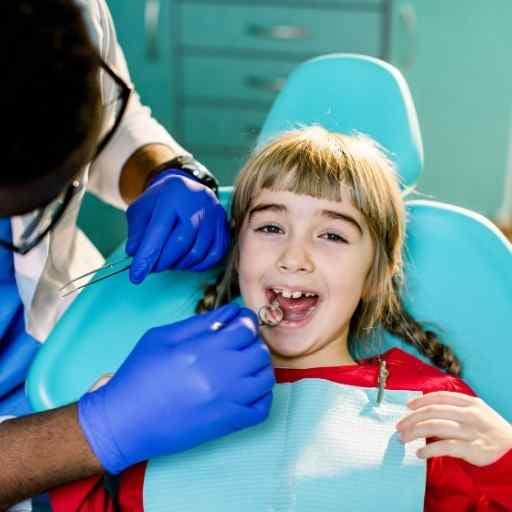 A young girl sits in a dental chair while a dentist examines her teeth with a dental tool.
